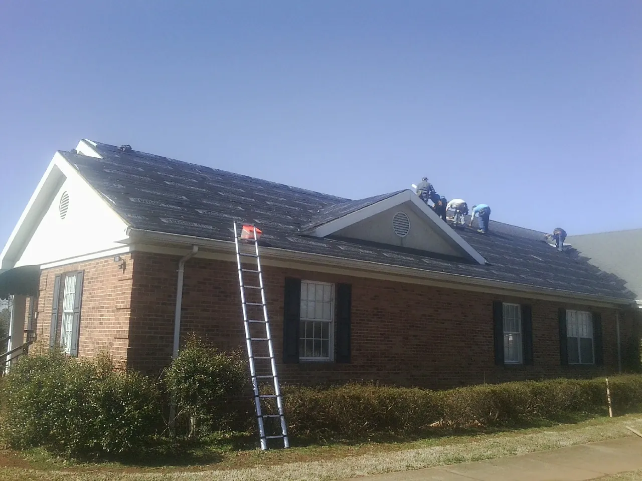 Crew tearing shingles off a brick ranch roof with a ladder set — Metal Roof Repair work by our crew in Balsam