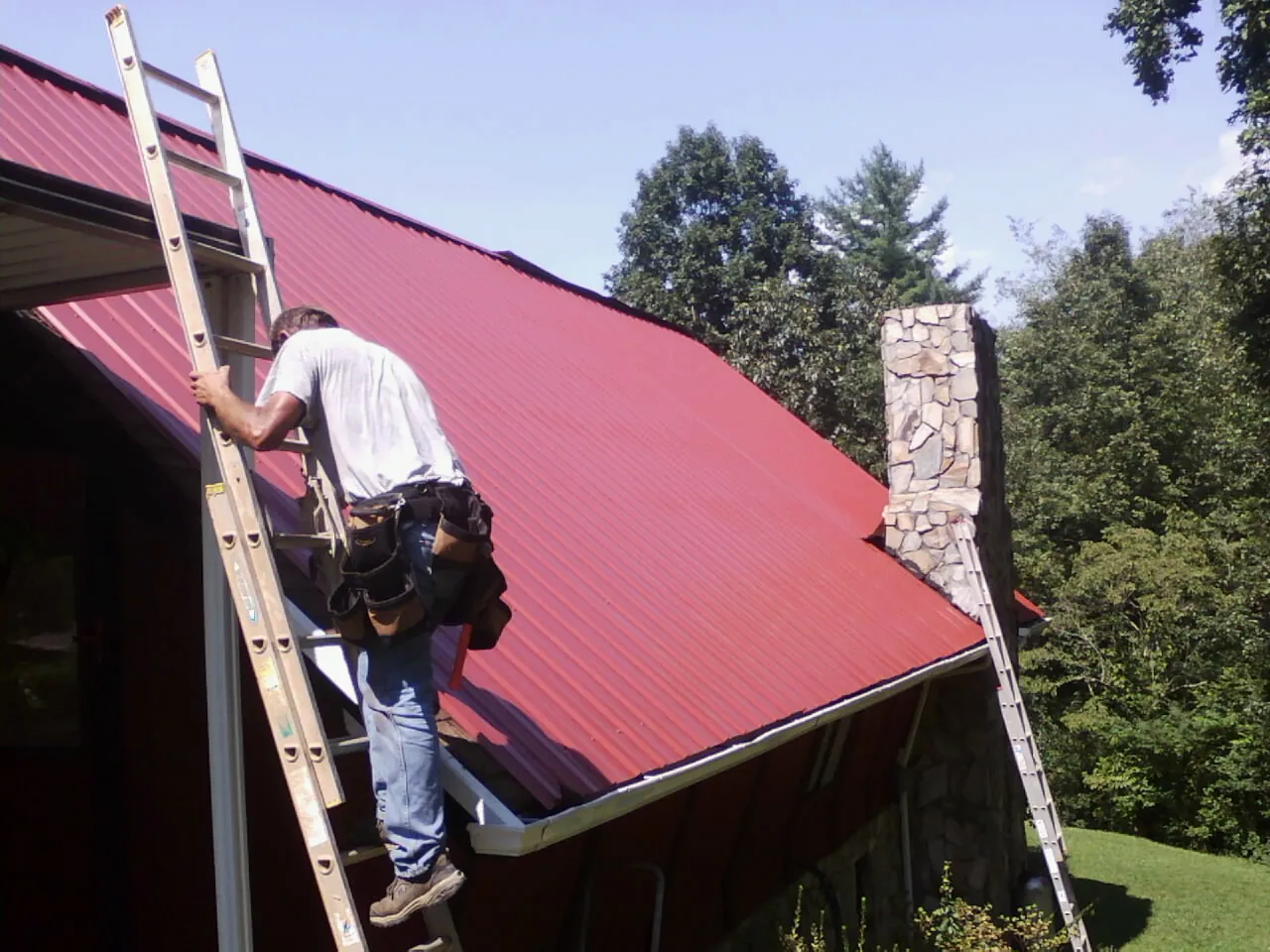 Roofer on a ladder beside a red metal roof and stone chimney — Soffit & Fascia Repair by Roofing Brothers in Balsam